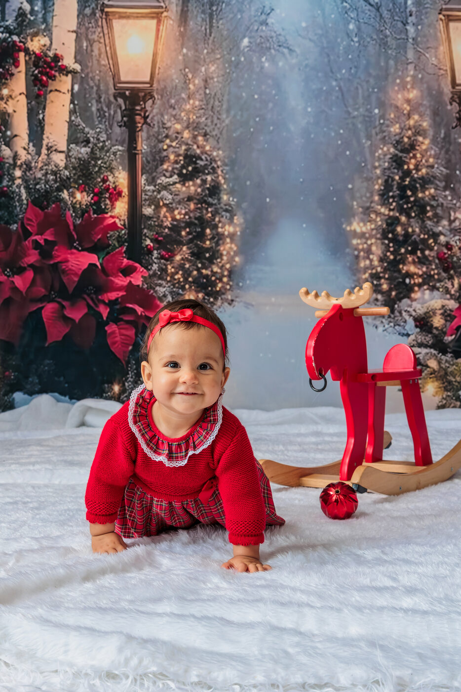 Bebé de vermelho na neve com uma rena de madeira, Natal de 2025